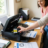 A woman in a home office smiles as she inserts the yellow Balloon Brand LC103 ink cartridge into a printer, with another woman looking on. Natural light from a window illuminates the scene.