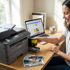 A smiling woman in a sunlit home office inserts the yellow Balloon Brand LC401XL ink cartridge into a Brother printer, next to her laptop and printed photos.