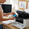 A woman in a home office with natural light is inserting the Balloon Brand Cyan ink cartridge into a Brother printer.