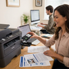 A woman in a home office is shown inserting a black Balloon Brand LC401XL ink cartridge into a Brother printer. The scene is bright and natural , with other people working in the background.